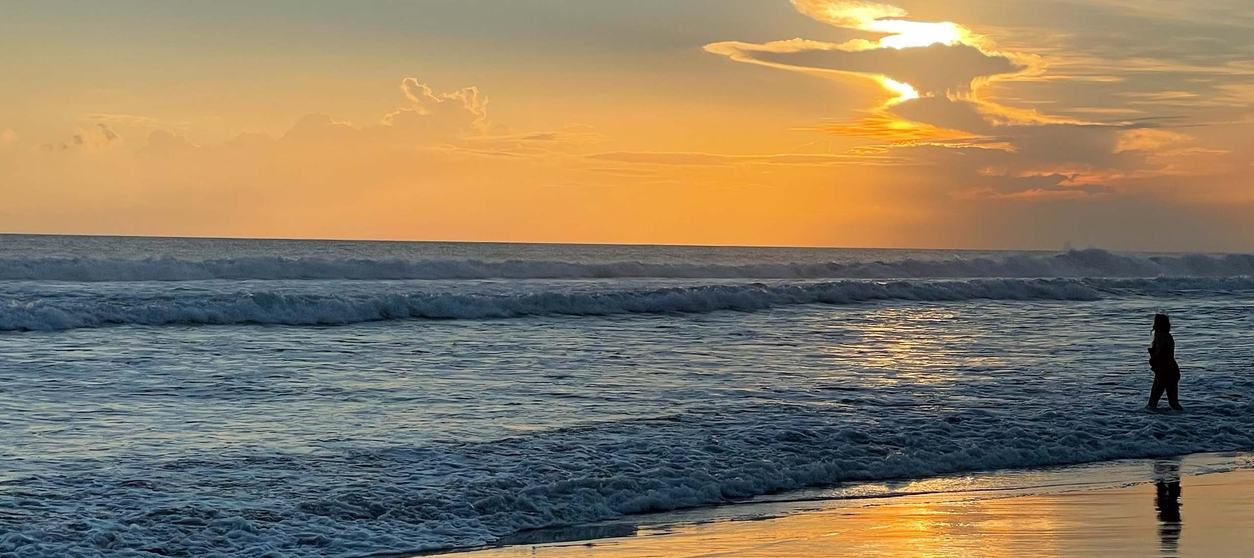 A lone figure standing in the shallows at sunset on a wide ocean beach